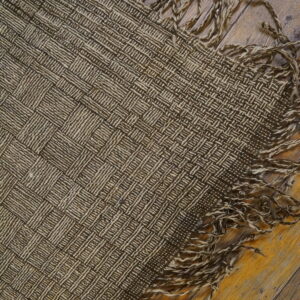 Close-up of a dark brown and tan basketweave rug with loose, twisted fringe on rustic wooden flooring.