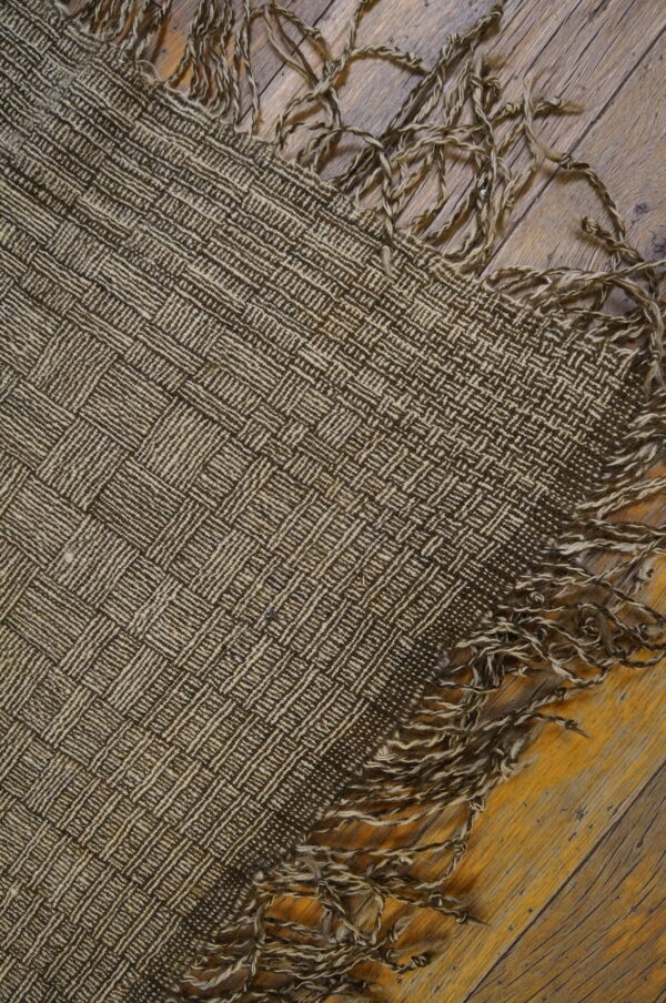 Close-up of a dark brown and tan basketweave rug with loose, twisted fringe on rustic wooden flooring.