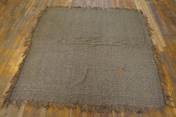 Worn green-brown flatweave rug with a checkerboard pattern and fringe rests on distressed wooden planks.