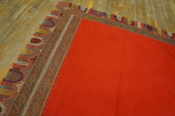 Solid orange rug field detailed with a complex paisley border resting on a distressed wood floor.