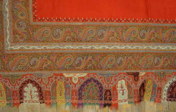 Close-up of a vermillion textile with multicolored paisley and arched mihrab motifs lying on distressed wood planks.