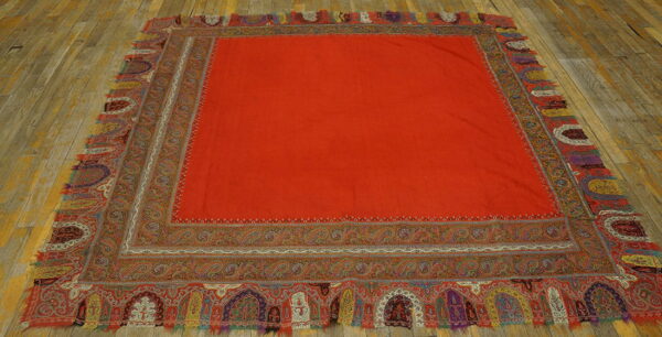 Square red flatweave textile rug with detailed multicolored paisley and arched borders on wood flooring.