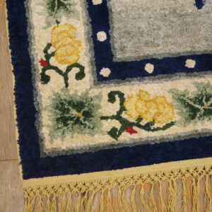 Highly textured floral rug with yellow blooms, a navy border, and long gold fringe on wood planks.