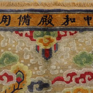 Ornate multicolor rug detail with sculpted pile, shimmering gold accents, and an orange border displaying black characters.