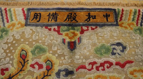 Ornate multicolor rug detail with sculpted pile, shimmering gold accents, and an orange border displaying black characters.