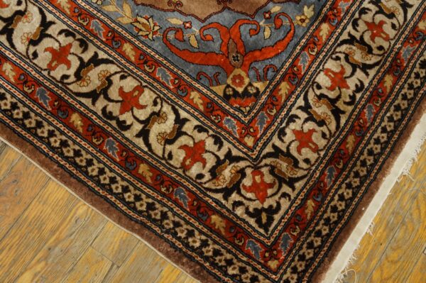 Ornate rug corner showing brown, blue, beige, and orange traditional patterns resting on light wood floors.