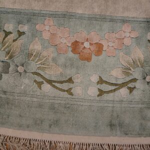 Sculpted floral rug border in sage green, pink, and tan with visible fringe lying on wood flooring.