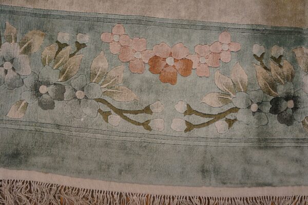 Vintage light cream color european influenced florals luxurious silk pile chinese room size rug 6 Sculpted floral rug border in sage green, pink, and tan with visible fringe lying on wood flooring.