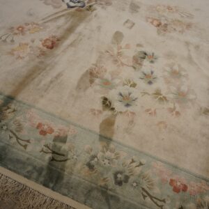 Muted floral rug with a carved cream field and teal border displays soft pile and fringes on dark wood.