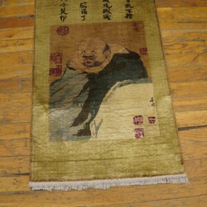 Shaggy olive green and brown runner rug depicting a laughing figure and calligraphy on worn wood floors.