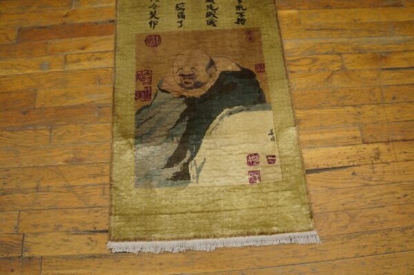Shaggy olive green and brown runner rug depicting a laughing figure and calligraphy on worn wood floors.
