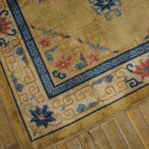 Gold rug with detailed blue and pink floral border lies on distressed yellow-toned wood flooring.