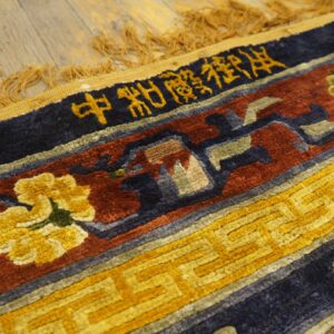 Textured rug border in navy, red, and gold, with script and floral designs, lies on wood planks.