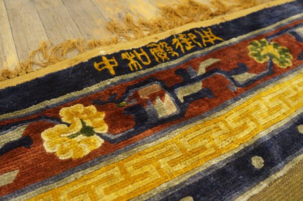 Textured rug border in navy, red, and gold, with script and floral designs, lies on wood planks.