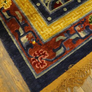 A high-pile rug with blue, red, and yellow floral patterns rests on worn wood planks, showing gold fringe.