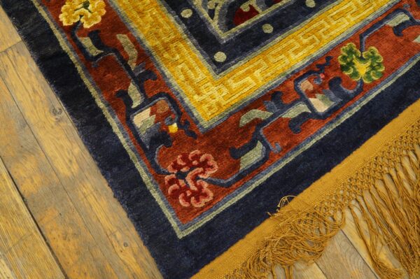 A high-pile rug with blue, red, and yellow floral patterns rests on worn wood planks, showing gold fringe.
