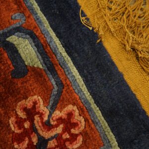 Textured rug detail in rust and navy borders a gold flatweave edge with thick fringe on wood flooring.