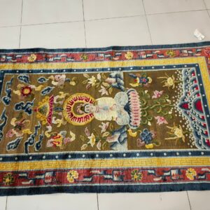 Brown rug with a central stylized figure and complex red, yellow, and blue borders on a white tile floor.