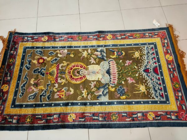 Brown rug with a central stylized figure and complex red, yellow, and blue borders on a white tile floor.