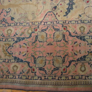 Ornate rug detail featuring pink, beige, and dark blue floral motifs, lying on wood flooring.