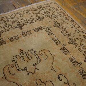 Low-pile beige rug with dark brown patterned border details rests on worn hardwood flooring.