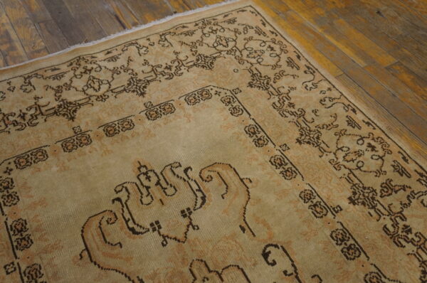Low-pile beige rug with dark brown patterned border details rests on worn hardwood flooring.
