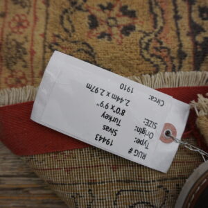 A tan and rust patterned rug with fringes is folded on wooden planks, partially covered by a product tag.