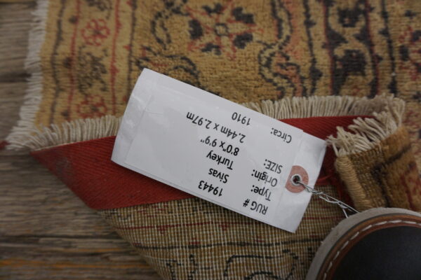 A tan and rust patterned rug with fringes is folded on wooden planks, partially covered by a product tag.