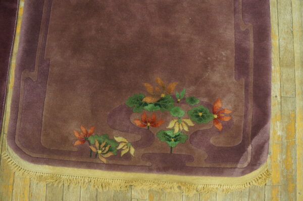 Close-up of a plum rug with orange and green floral carvings and cream fringe on distressed wooden floorboards.