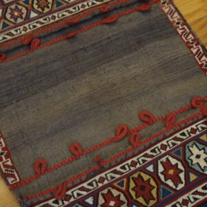 Geometric patterned flatweave textile folded over a plain gray panel accented with decorative red braided trim.