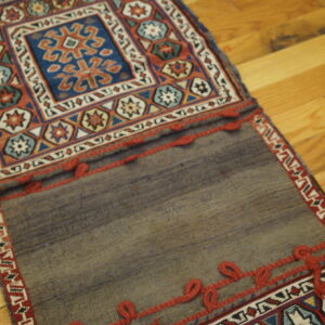 Multicolored geometric patterns in blue and rust contrast with a plain gray flatweave, both trimmed with red loops on wood.