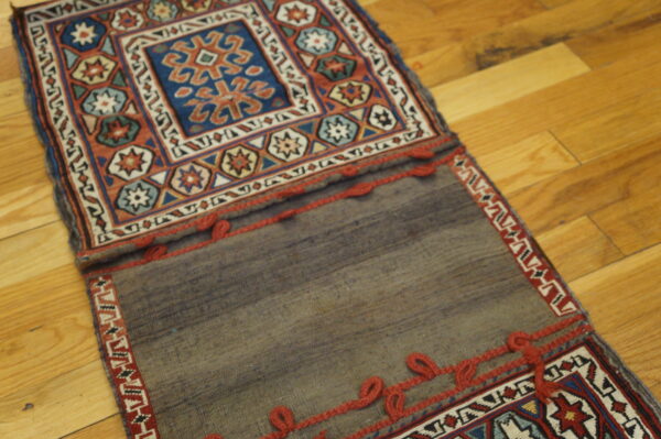 Multicolored geometric patterns in blue and rust contrast with a plain gray flatweave, both trimmed with red loops on wood.