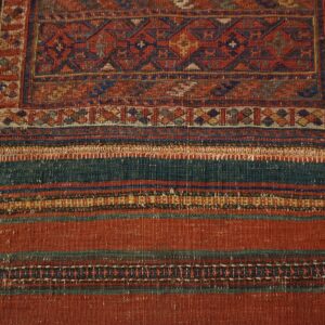 Woven textile with a geometric red and blue border above wide flatweave stripes on weathered wood.