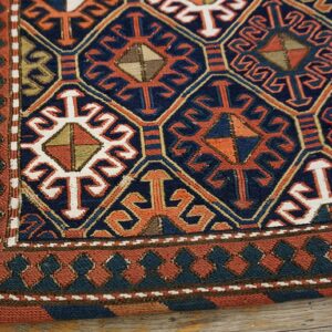Navy and orange geometric rug with stylized motifs and patterned borders resting on wood floorboards.