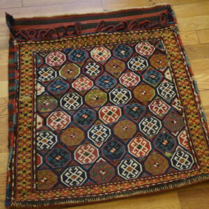 Small textile displaying dark blue, red, and green geometric patterns with looped fringe detail on hardwood.