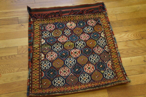 Small textile displaying dark blue, red, and green geometric patterns with looped fringe detail on hardwood.