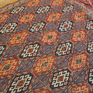 Intricate geometric diamond rug pattern in red, navy blue, and gold resting on wooden flooring.