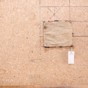 A small, plain beige rug sample with multicolor fringe sits on marked chipboard next to wood plank flooring.