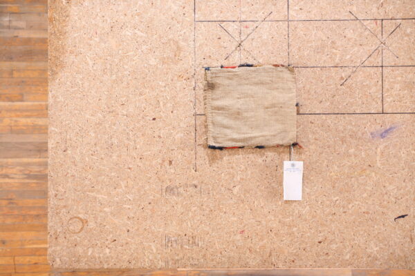 A small, plain beige rug sample with multicolor fringe sits on marked chipboard next to wood plank flooring.