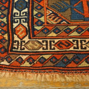 Flatwoven rug corner with dense geometric patterns in orange, blue, and beige, lying on wood planks.