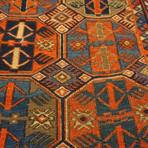 Close-up of a low-pile rug featuring a dense geometric pattern in orange-red, navy, teal, and brown.
