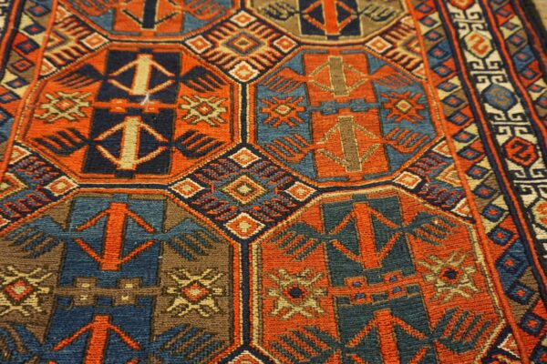 Close-up of a low-pile rug featuring a dense geometric pattern in orange-red, navy, teal, and brown.
