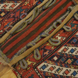 Red and black patterned rug fragment with a striped panel, braided loops, and tan leather strips on a wood floor.