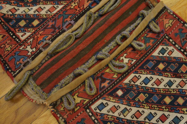 Red and black patterned rug fragment with a striped panel, braided loops, and tan leather strips on a wood floor.
