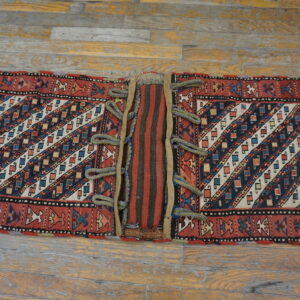 Red, blue, and cream geometrically patterned textile with a striped center panel and corded loops on wooden planks.