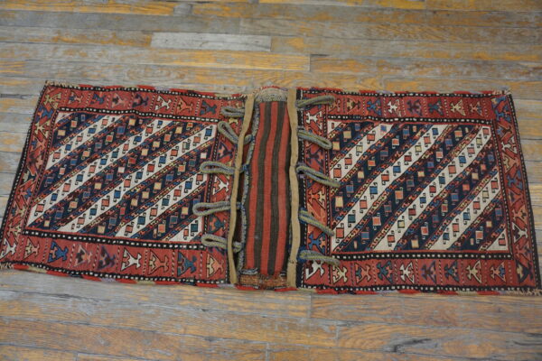 Red, blue, and cream geometrically patterned textile with a striped center panel and corded loops on wooden planks.