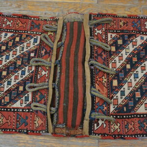A flatwoven red and blue saddlebag face with diagonal geometric stripes and braided loops, resting on wood floorboards.