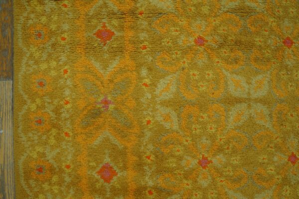 Textured, golden yellow rug with orange and pink abstract patterns next to wooden flooring.