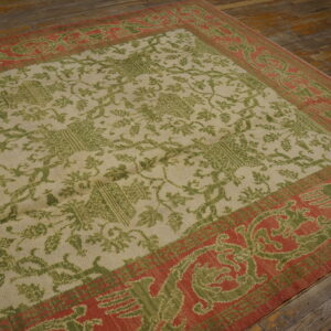 Beige rug with a green floral pattern and a wide red border on rustic wooden floorboards.