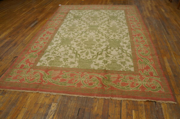 A low-pile rug with a cream field, green vine pattern, and a coral-and-green ornate border on wood flooring.